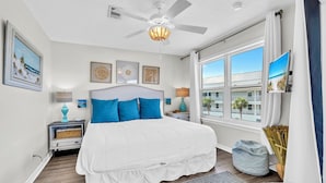 A bedroom with a large bed, a ceiling fan, a window with a view of buildings, and a TV on the wall.