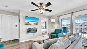 Een moderne woonkamer met een grijze bank, een blauwgroene fauteuil en een ronde salontafel. Een plafondventilator, een flatscreen-televisie en een lamp zijn zichtbaar. De kamer heeft een groot raam met uitzicht op een balkon en gebouwen.
