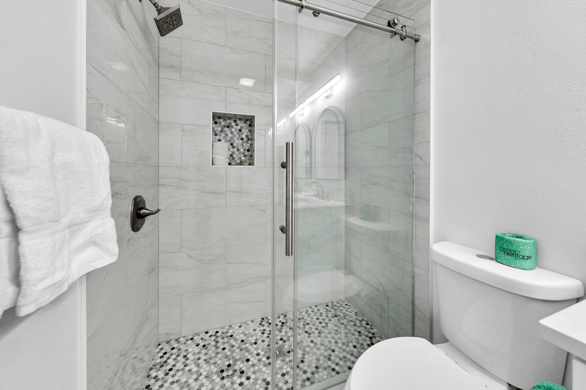 A modern bathroom with a glass-enclosed shower, a toilet with a green tissue box, and a patterned floor.