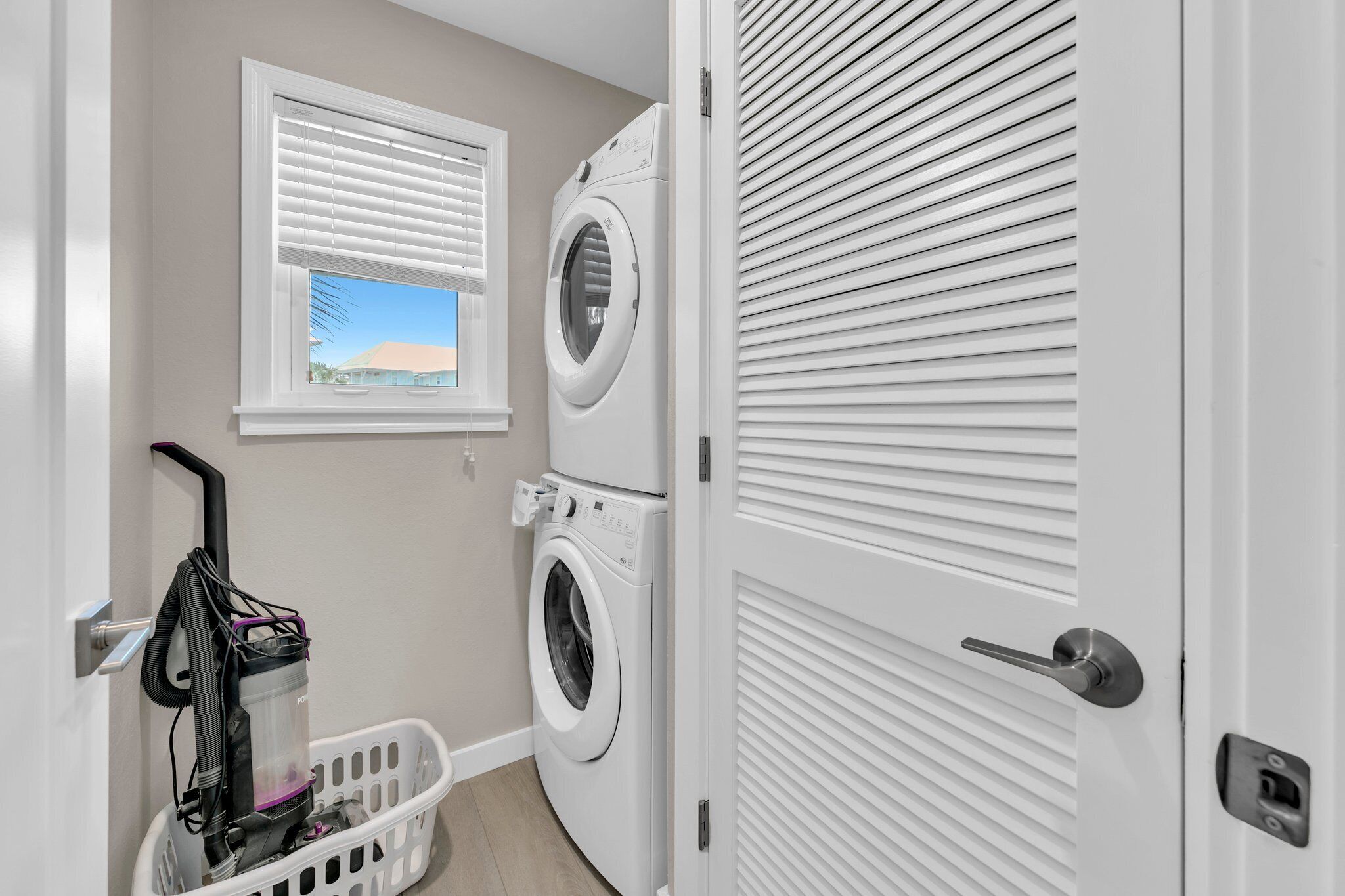 A laundry room with a front-loading washer and dryer, a window with blinds, and a door with a louvered design.
