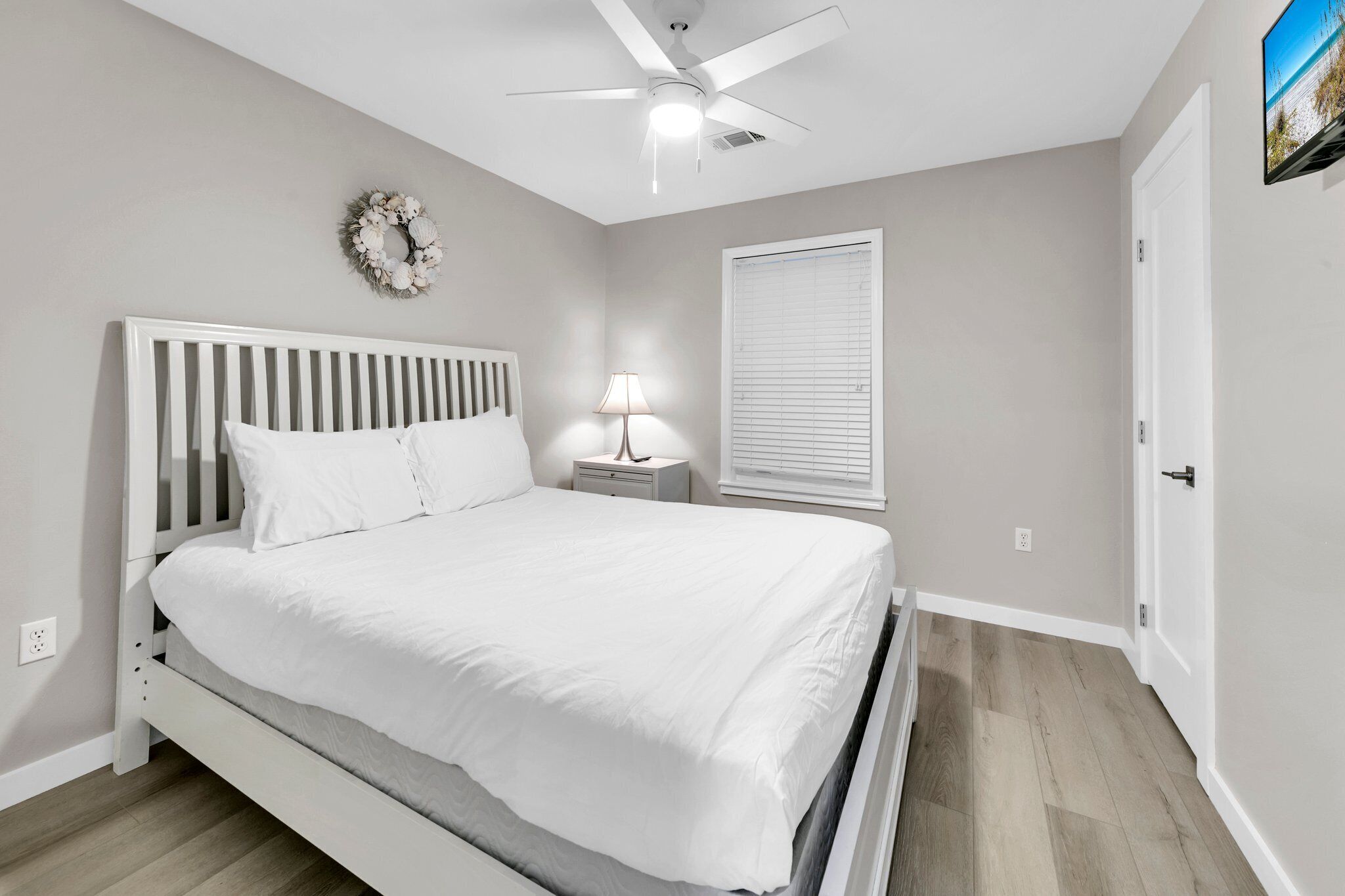 Una camera da letto con un letto bianco, una ventola a soffitto, un comodino con una lampada, una finestra con persiane e una porta.