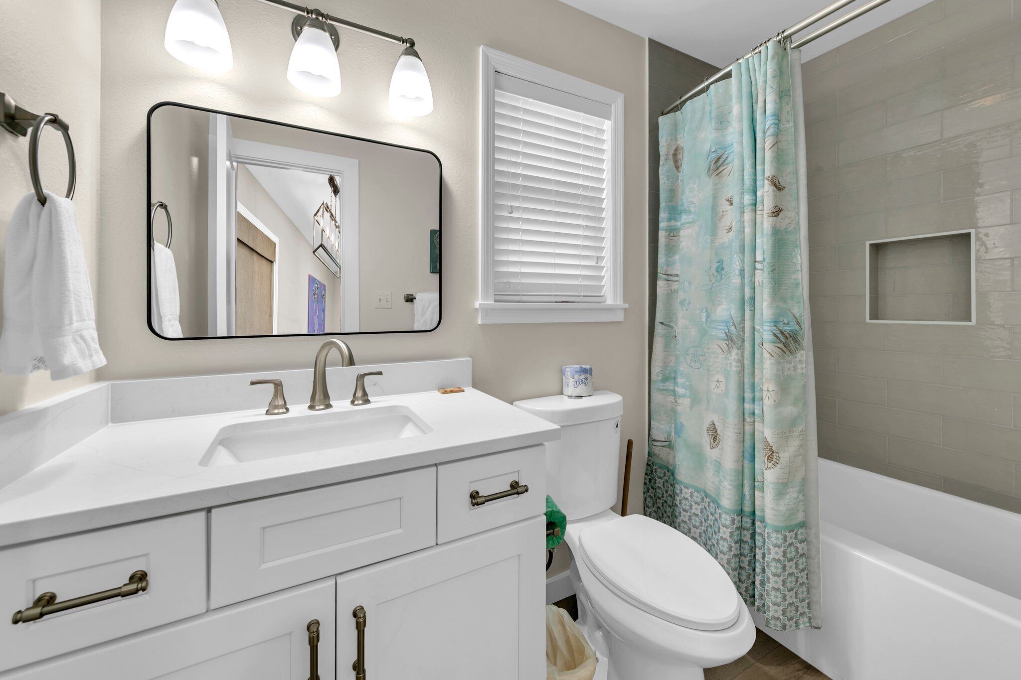 A bathroom with a white sink, mirror, and toilet, a shower curtain, and a window with blinds.