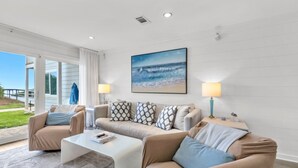 Un salon avec un canapé beige, des coussins à motifs, une table basse blanche et un tableau sur le mur représentant un paysage de plage.