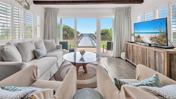 Amplia sala de estar con televisor grande, asientos cómodos y vista a la playa a través de ventanas amplias.