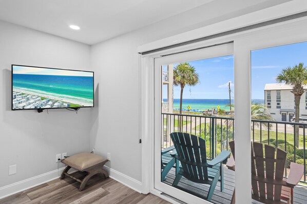 A balcony with a view of the ocean and beach, a television mounted on the wall, and a bench.