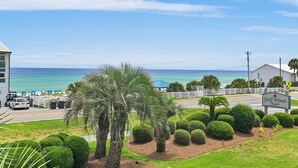 A view of a coastal area with a sign for 'Gulf Winds East' hotel, palm trees, neatly trimmed bushes, and a clear blue sky. - ⭐Beach Views -Gated Community Pool - Balconies-2BR Gulf Winds East #3 ⭐ (Miramar Beach)