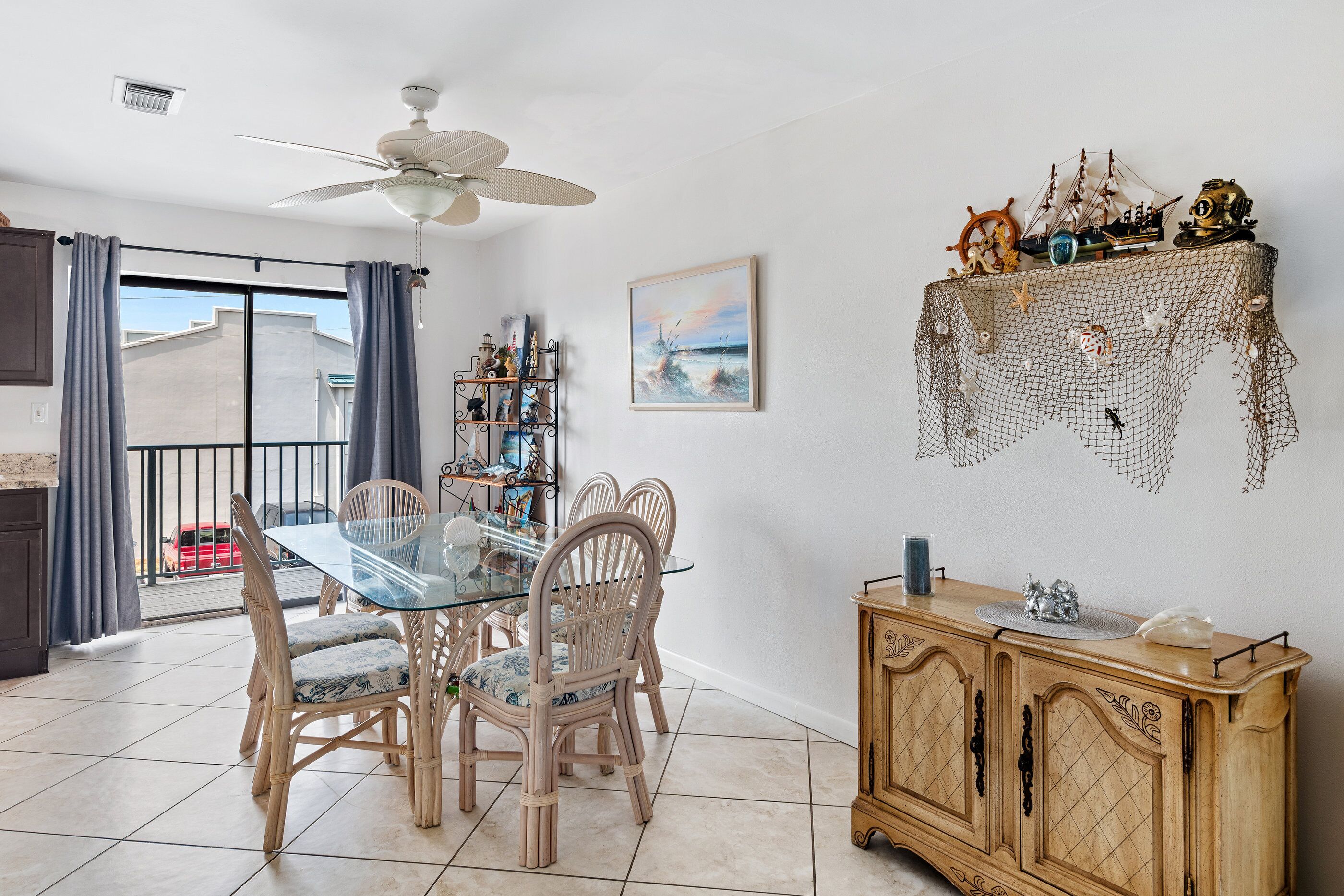 Un comedor con mesa de vidrio y sillas de mimbre, un armario de madera con objetos decorativos, y vistas al exterior a través de una puerta corrediza.