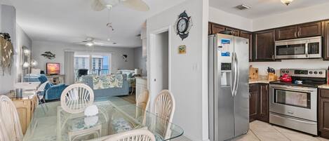 A spacious kitchen with modern appliances, a dining area with glass-top table and wicker chairs, and a ceiling fan.