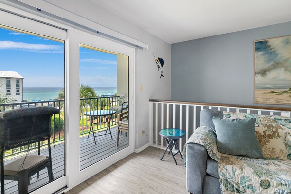 A balcony with a view of the ocean, a dining area with chairs and a table, and a living area with a sofa and cushions.