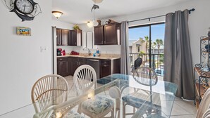 A dining area with a glass table and chairs, a kitchen with dark cabinets, a decorative clock, and a view of the outside. - ⭐Beach Views -Gated Community Pool - Balconies-2BR Gulf Winds East #3 ⭐ (Miramar Beach)