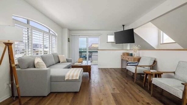 Una sala de estar moderna con un sofá seccional gris, piso de madera, un televisor montado en la pared y vistas al exterior a través de amplios ventanales.