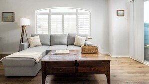 Una sala de estar con un sofá gris, una mesa de centro de madera y un ventanal con contraventanas blancas.