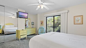 Un dormitorio con cama, ventilador de techo, televisor, cómoda, silla y balcón con vistas a árboles.
