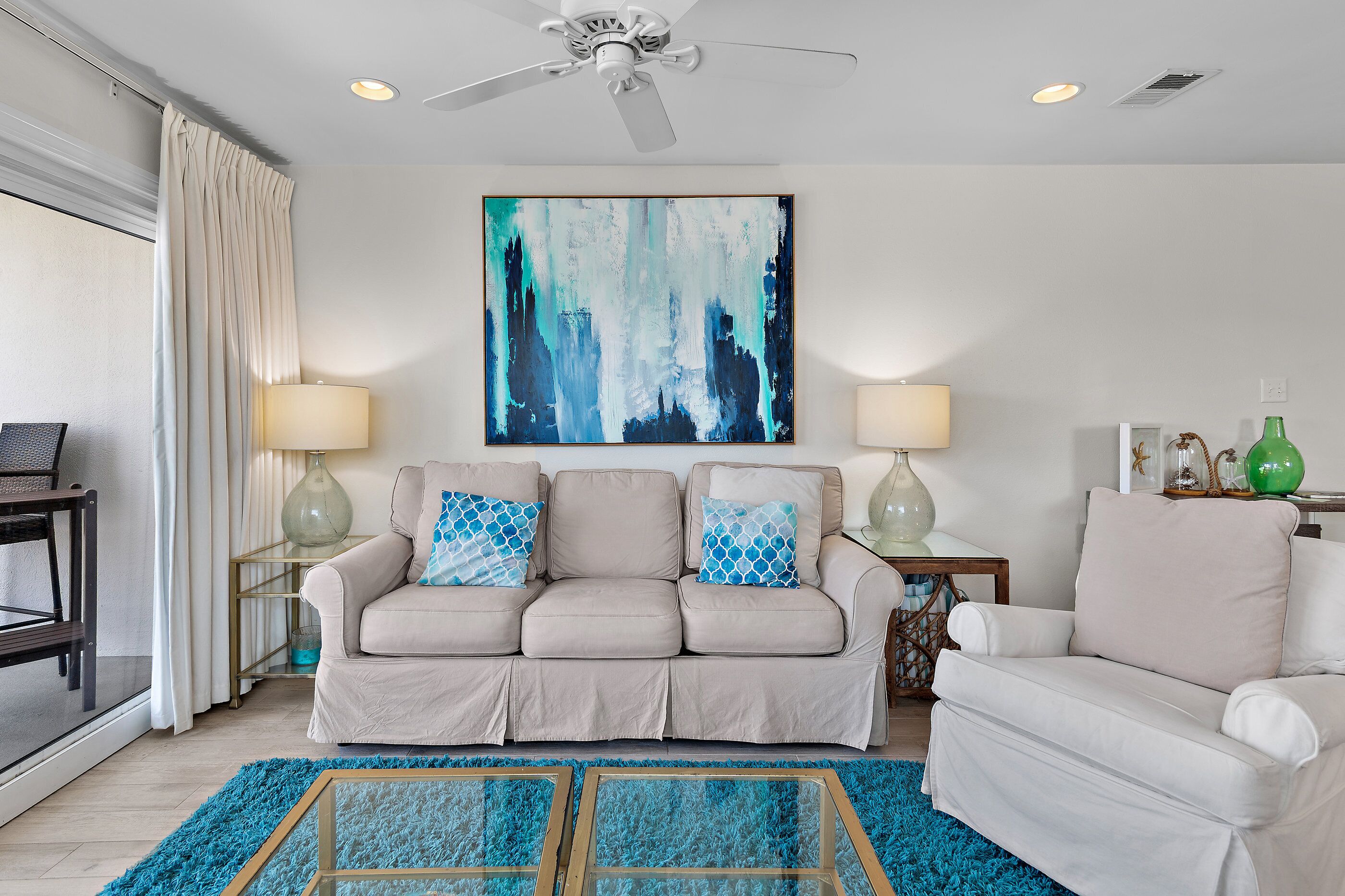 A living room with a beige sofa, two side tables with lamps, a large abstract painting, and a ceiling fan.