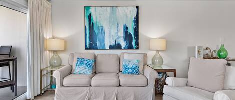 A living room with a beige sofa, two side tables with lamps, a large abstract painting, and a ceiling fan.