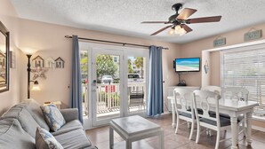 A living room with a grey sofa, white coffee table, and a dining area with a white table and chairs. There is a ceiling fan, a TV, and a view of the outside through a sliding door.