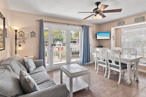 A living room with a grey sofa, white coffee table, and a dining area with a white table and chairs. There is a ceiling fan, a TV, and a view of the outside through a sliding door.