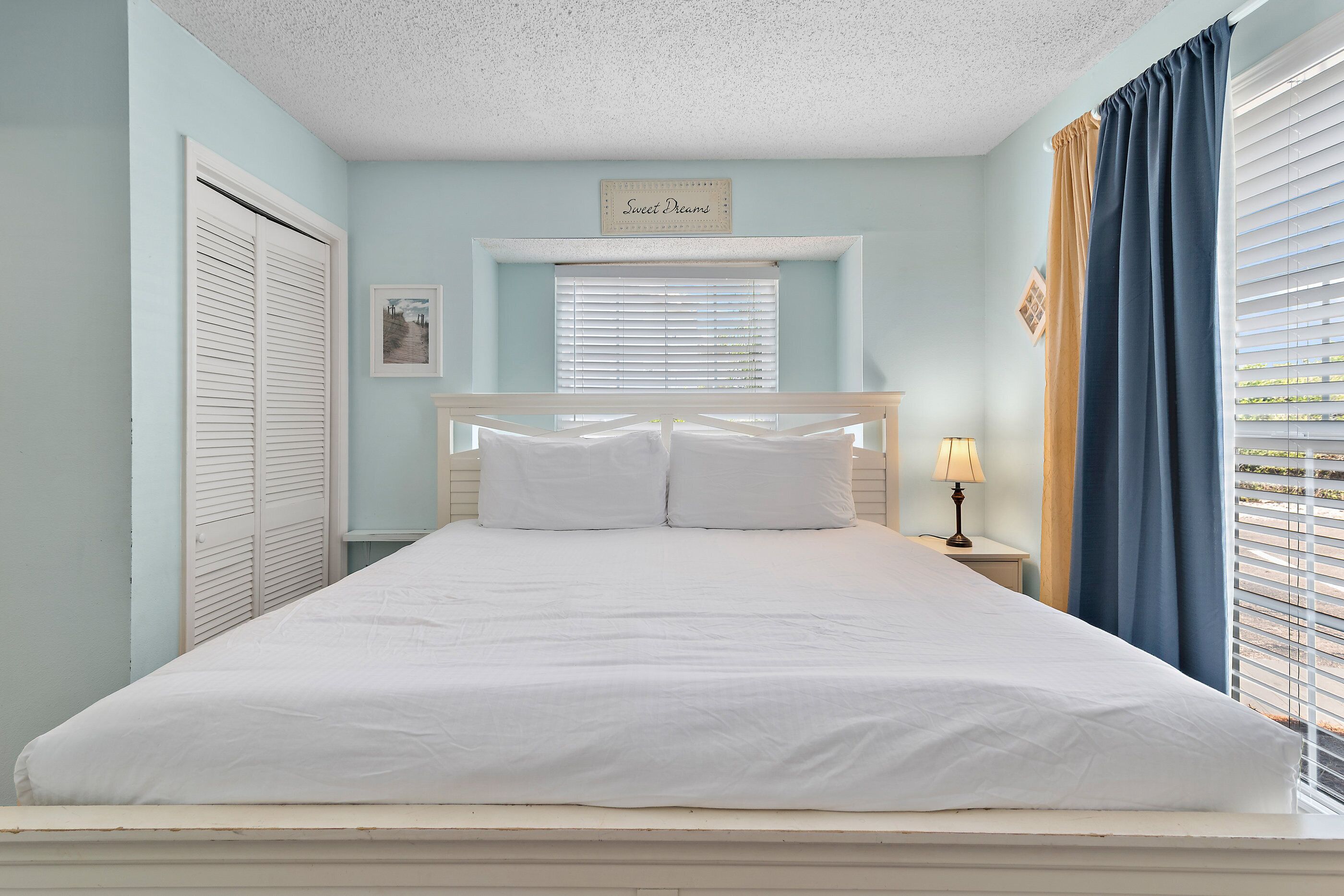 A bedroom with a large bed, a nightstand, a lamp, a window with blinds, and a sign above the window.