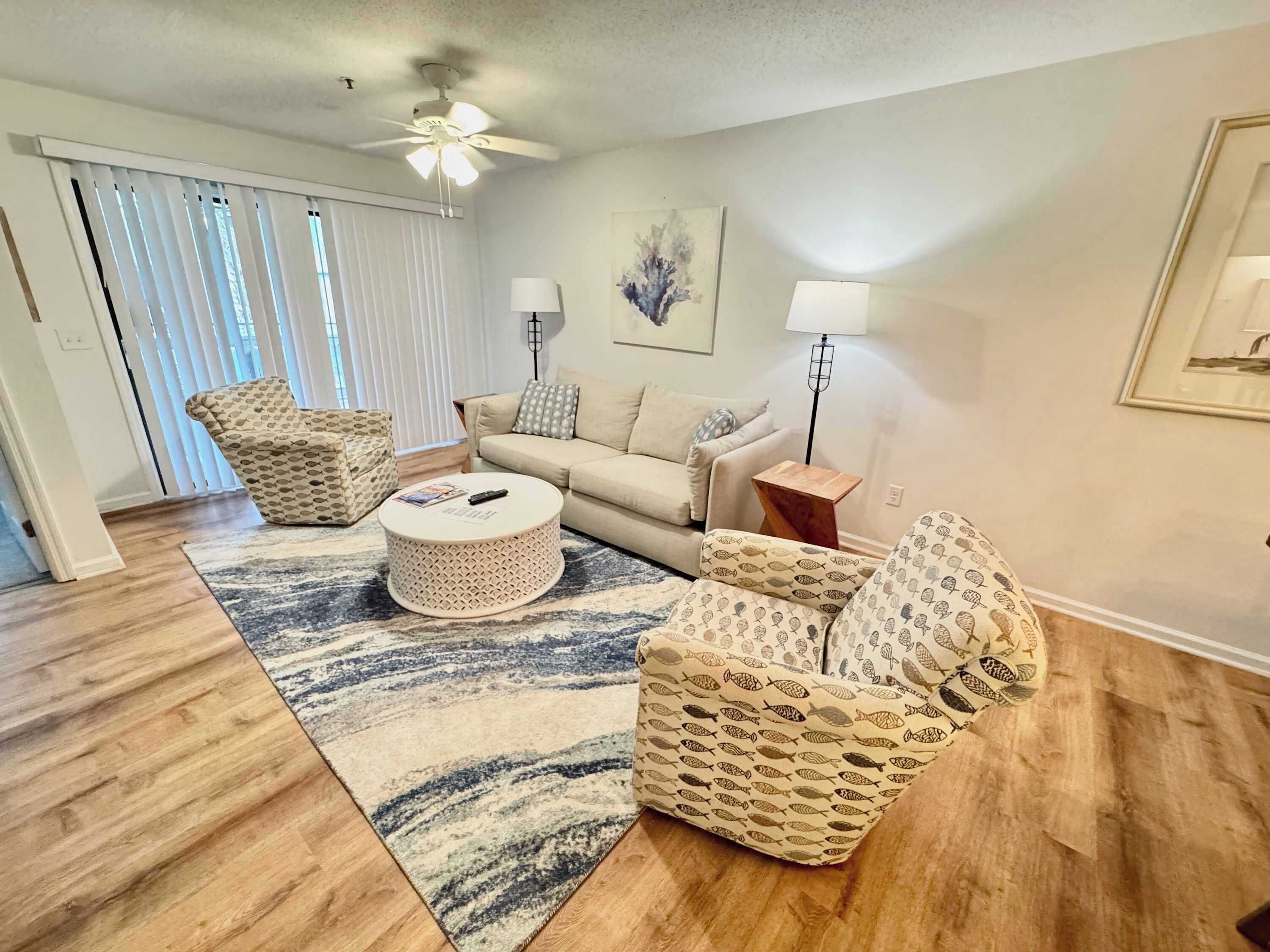 A living room with a sofa, two patterned armchairs, a coffee table, a floor lamp, and a ceiling fan.
