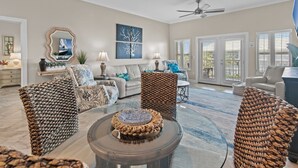 A spacious living room with a large round coffee table, a sofa, and a chair with a woven backrest. There is a ceiling fan, a painting on the wall, and a window with blinds.