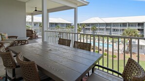 A covered outdoor dining area with a wooden table and wicker chairs, overlooking a swimming pool and a building with multiple balconies.