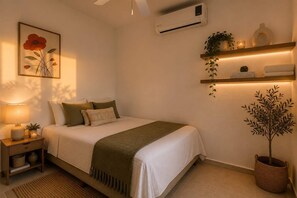 A neatly arranged bedroom with a bed, bedside table, lamp, wall-mounted air conditioner, and decorative items including a framed picture and shelves with plants.