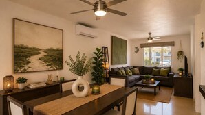 A modern living room with a dining area, featuring a sofa set, a coffee table, and a dining table with chairs. There is a large painting on the wall, a ceiling fan, and a window providing natural light. - Relax in Nuevo Vallarta | Pool & Great Location (Nuevo Vallarta)