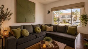 A living room with a sectional sofa, a wall-mounted abstract painting, a ceiling fan, a large window with a view of the outside, and a small side table with a plant and a candle. - Relax in Nuevo Vallarta | Pool & Great Location (Nuevo Vallarta)