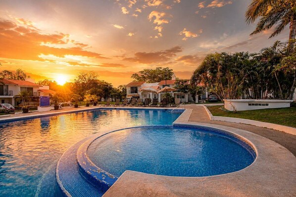 A swimming pool with a curved edge, surrounded by lounge chairs and palm trees, with a sunset in the background.