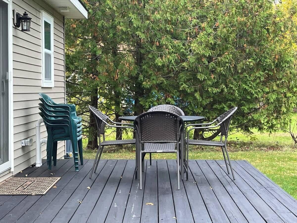 Una terraza de madera con una mesa y sillas, un porche con una puerta, y una casa al fondo.