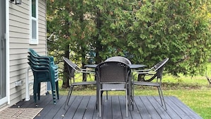 Um deck de madeira com mesa e cadeiras, um alpendre com porta e uma casa ao fundo.