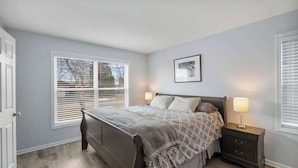 Quarto com cama, mesas de cabeceira, janela com persianas e um quadro na parede.
