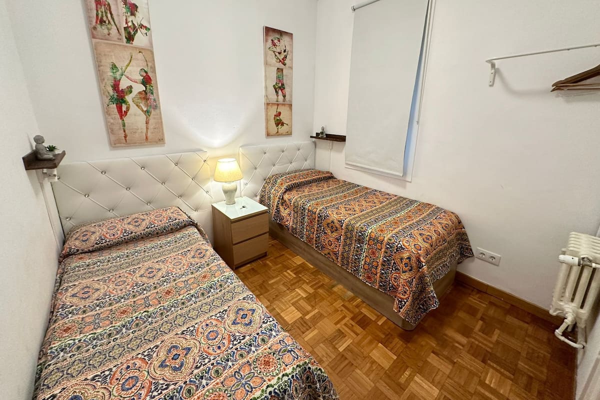 A small, clean room with two beds, a nightstand with a lamp, and artwork on the walls.