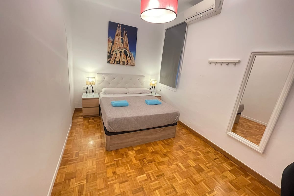 A bedroom with a bed, two bedside tables, a nightstand, a large mirror, and a painting of a cathedral.