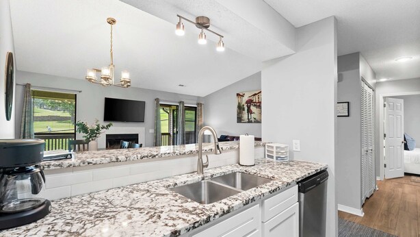 A kitchen with a granite countertop, stainless steel sink, and a dishwasher. There is a flat-screen TV mounted on the wall and a window with curtains. - Quittin' Time - Stay for 6 with Golf Course Views & Resort Amenities (Branson)