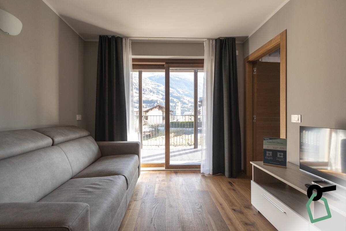 Una sala de estar moderna con un sofá, un televisor de pantalla plana y un puerta corrediza de vidrio que da a un balcón con vistas a la montaña.