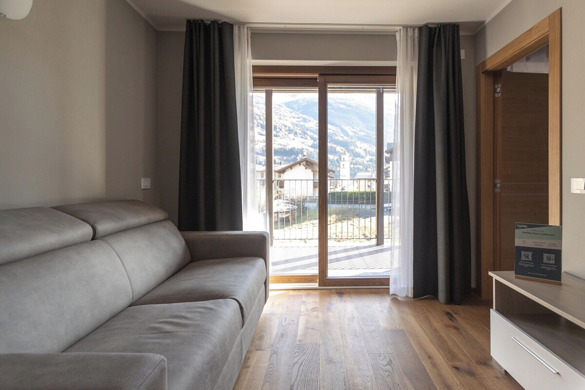 A modern living room with a sofa, a flat-screen TV, and a sliding glass door leading to a balcony with a mountain view.