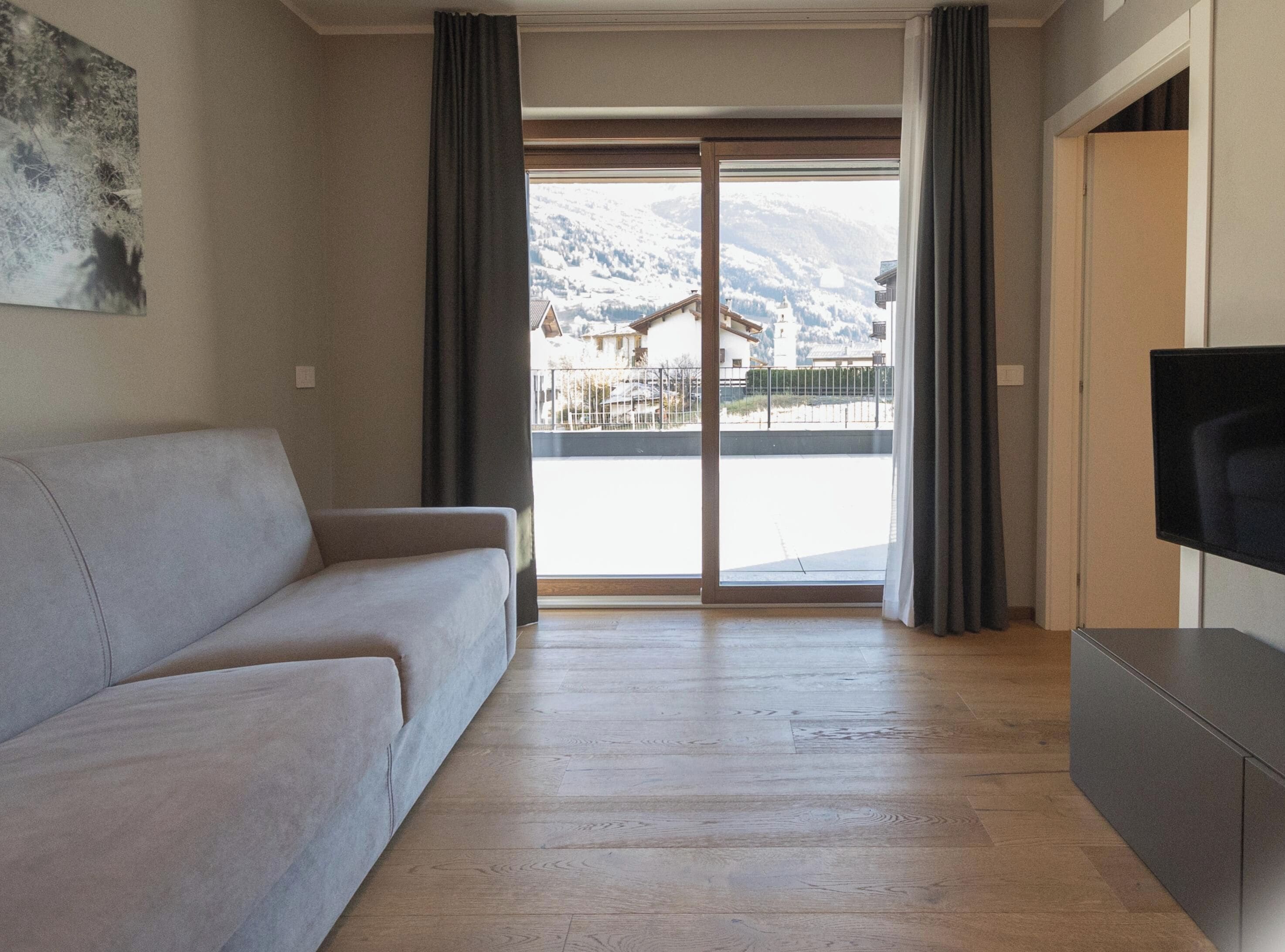 A living room with a sofa, a flat-screen TV, and a large sliding glass door leading to a balcony with a mountain view.