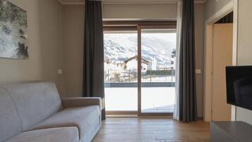 A living room with a sofa, a flat-screen TV, and a large sliding glass door leading to a balcony with a mountain view.