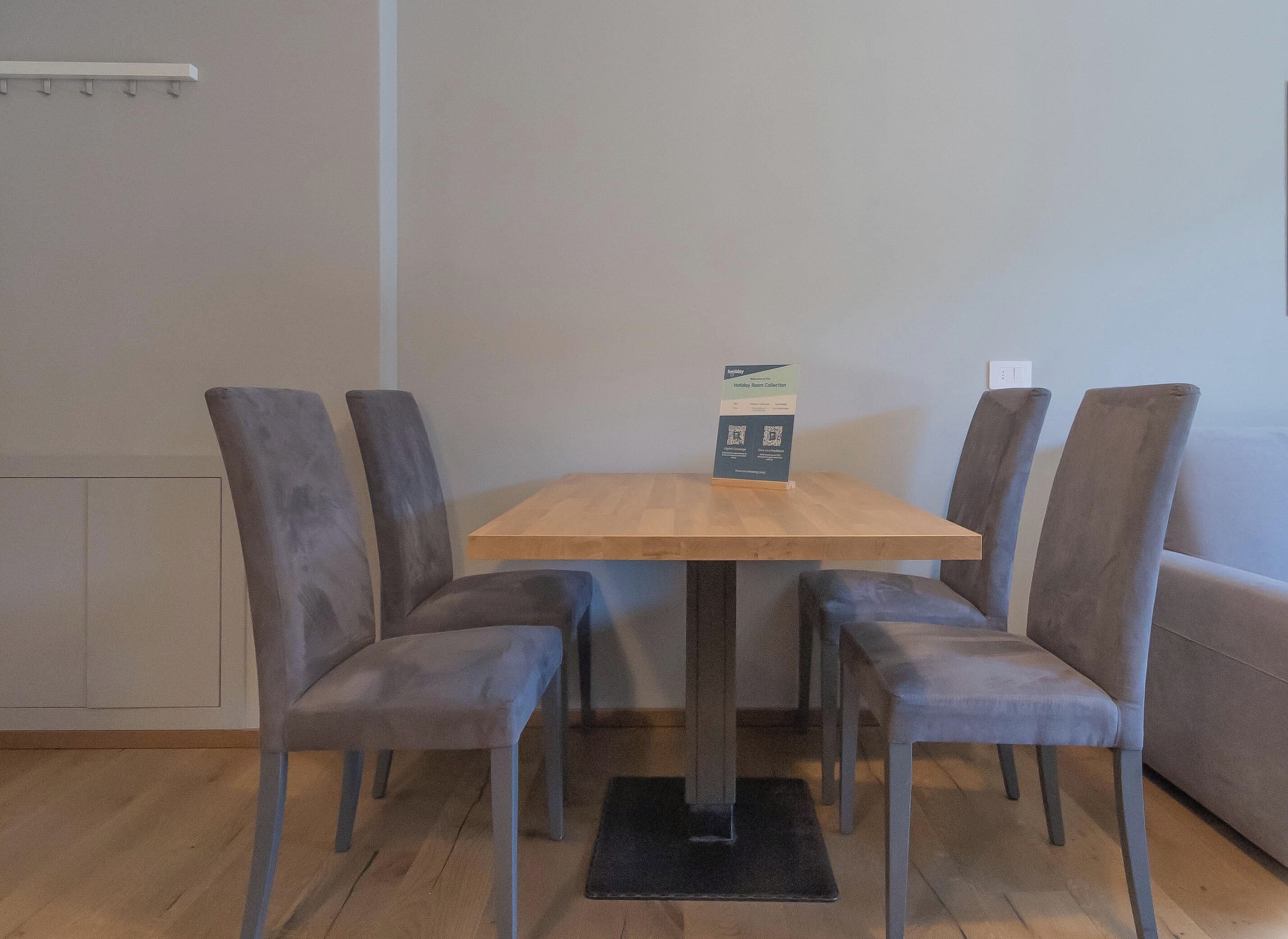 A dining area with a wooden table and four grey upholstered chairs. A sign on the table indicates a holiday room collection.