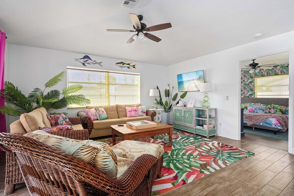 A living room with a sofa, wicker chairs, a coffee table, a patterned rug, a ceiling fan, and a bed in the adjacent room.