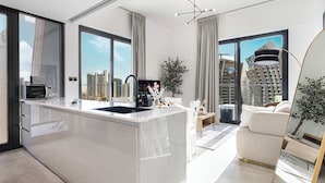 A modern kitchen with a central island, a dining area with a white table and chairs, and a living area with a sofa and a large window offering a city view. - Luxury 2BR with Canal View Burj Khalifa view and Infinity Pool Access (Business Bay)