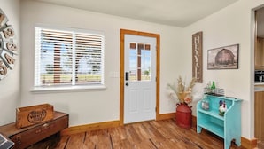 A room with a wooden floor, a white door, a window with blinds, a Red & White food stores sign, a rustic cabinet, and a teal shelf with bottles and a rooster figurine.