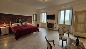 A bedroom with a large bed, bedside tables, a chair, a table with glasses and a bottle, and a TV on the wall.