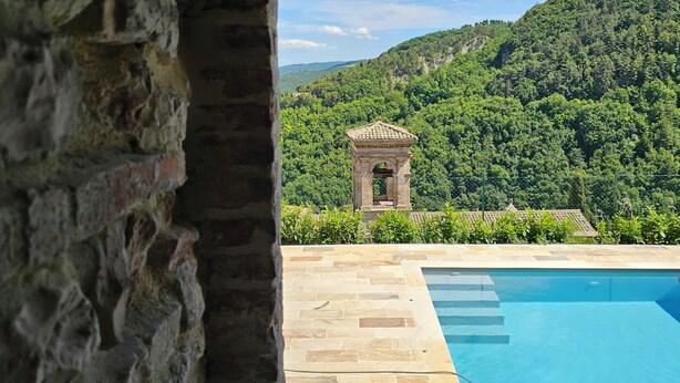 A stone archway leading to a swimming pool with a view of a lush green hillside and a small stone structure.