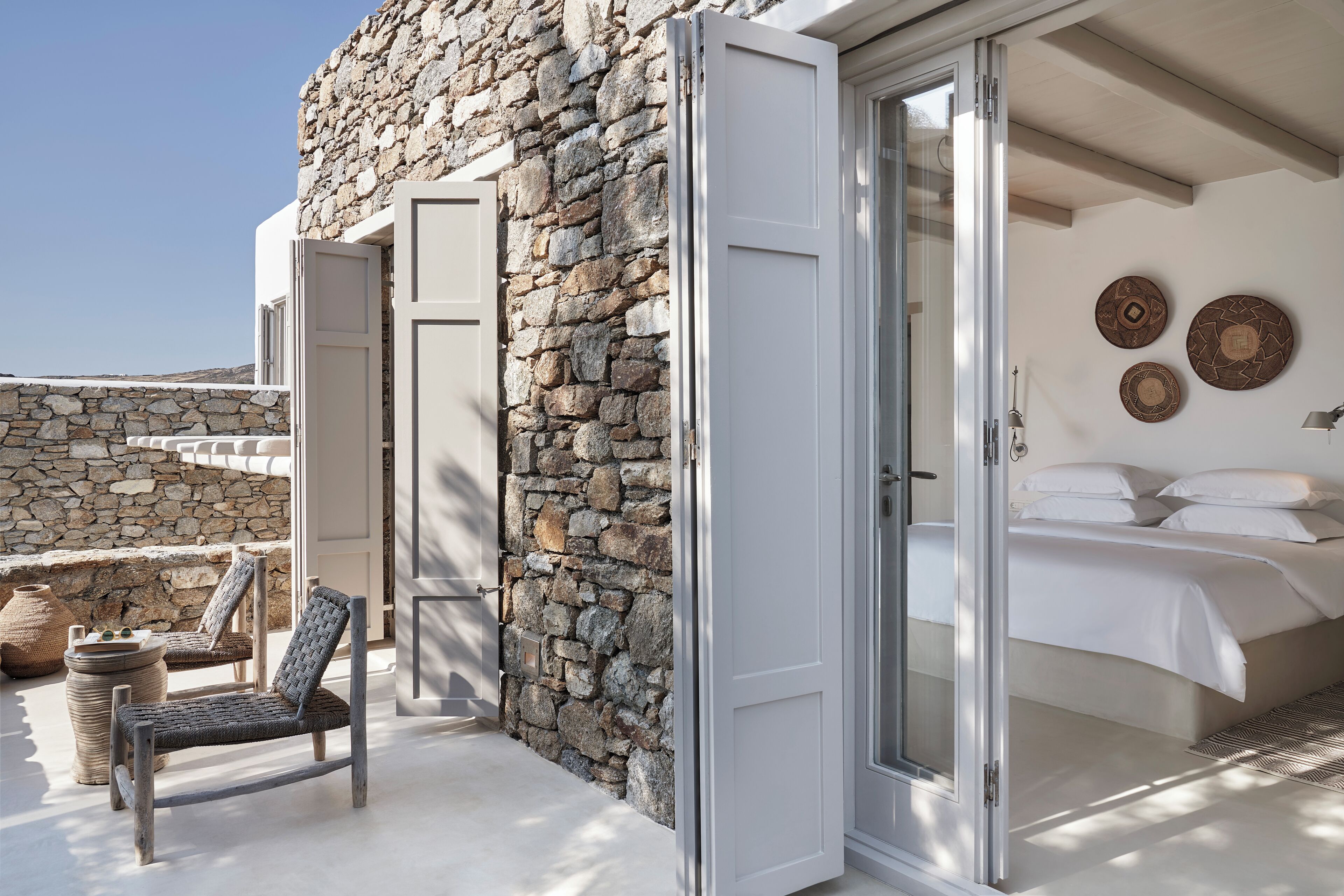 A bedroom with a stone wall, a bed with white linens, and a view of the outdoors.