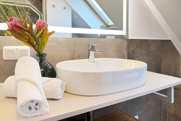 Un baño moderno con lavamanos blanco, un espejo y un florero con flores.