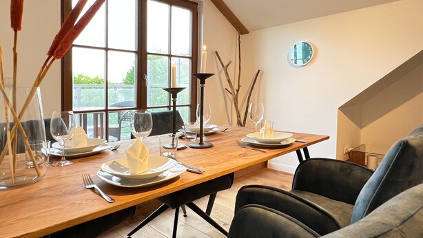 Een eetruimte met een houten tafel gedekt voor vier personen, omringd door stoelen, met een raam en een klok aan de muur.