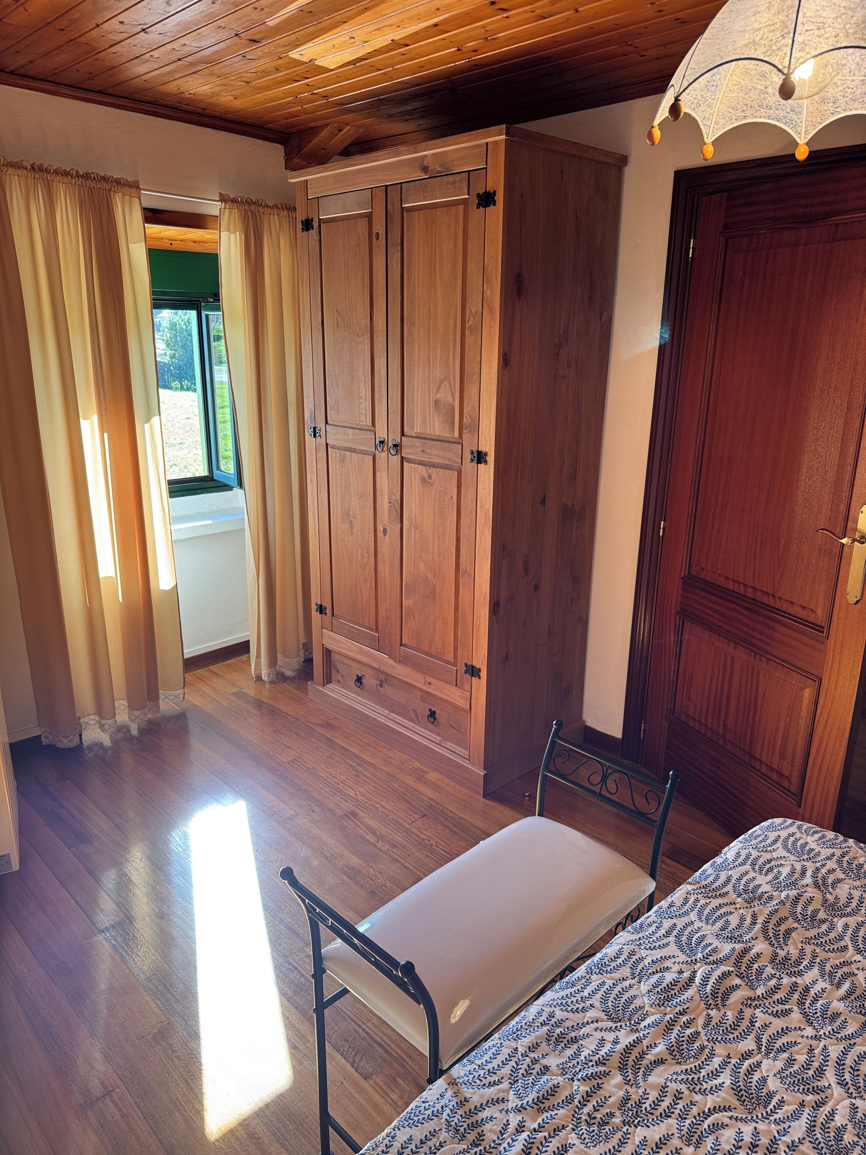 Una camera da letto con un'armadio in legno, un letto con copriletto decorato, una scrivania con un portatile e una finestra con tende.