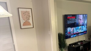 Een moderne woonkamer met een zwarte eettafel, twee zwarte stoelen, een flatscreen televisie aan de muur, een ingelijste kunstwerk en een potplant.